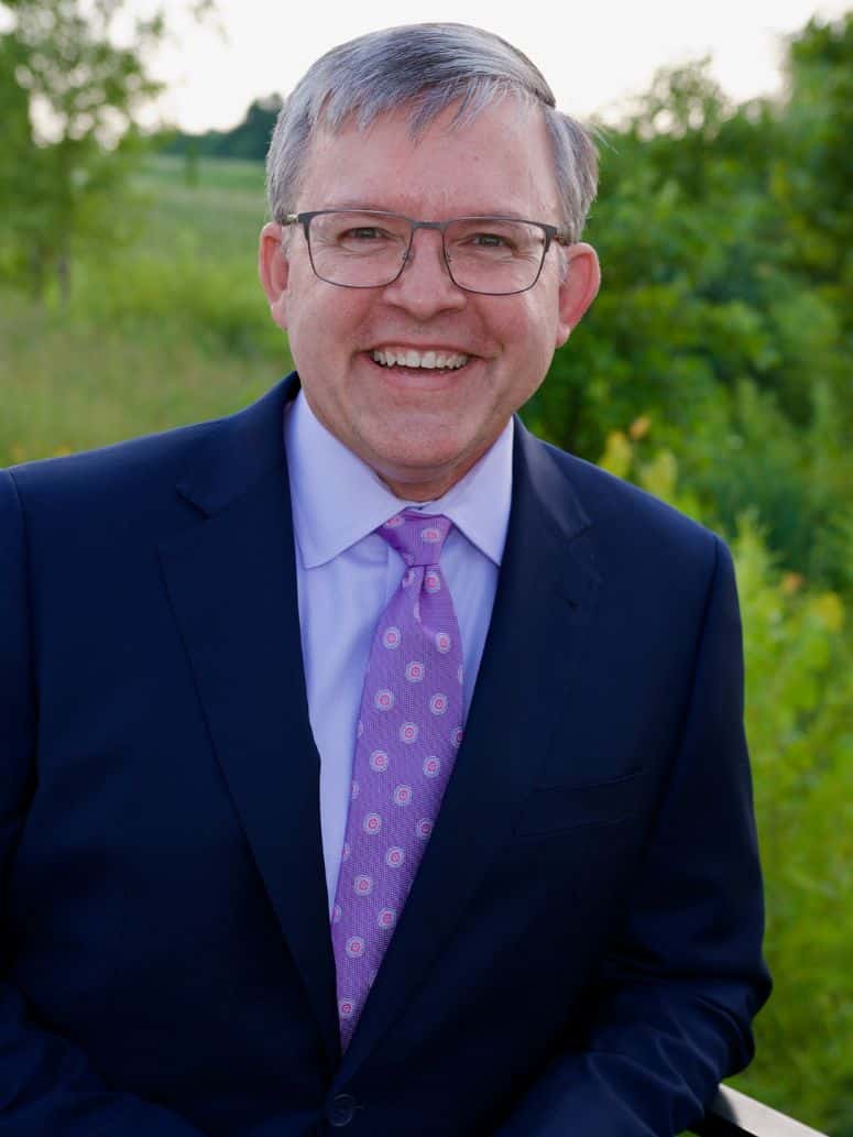 Blue Ash Council Member Brian Gath poses in a blue suit and purple shirt in front of a blurry green natural background.